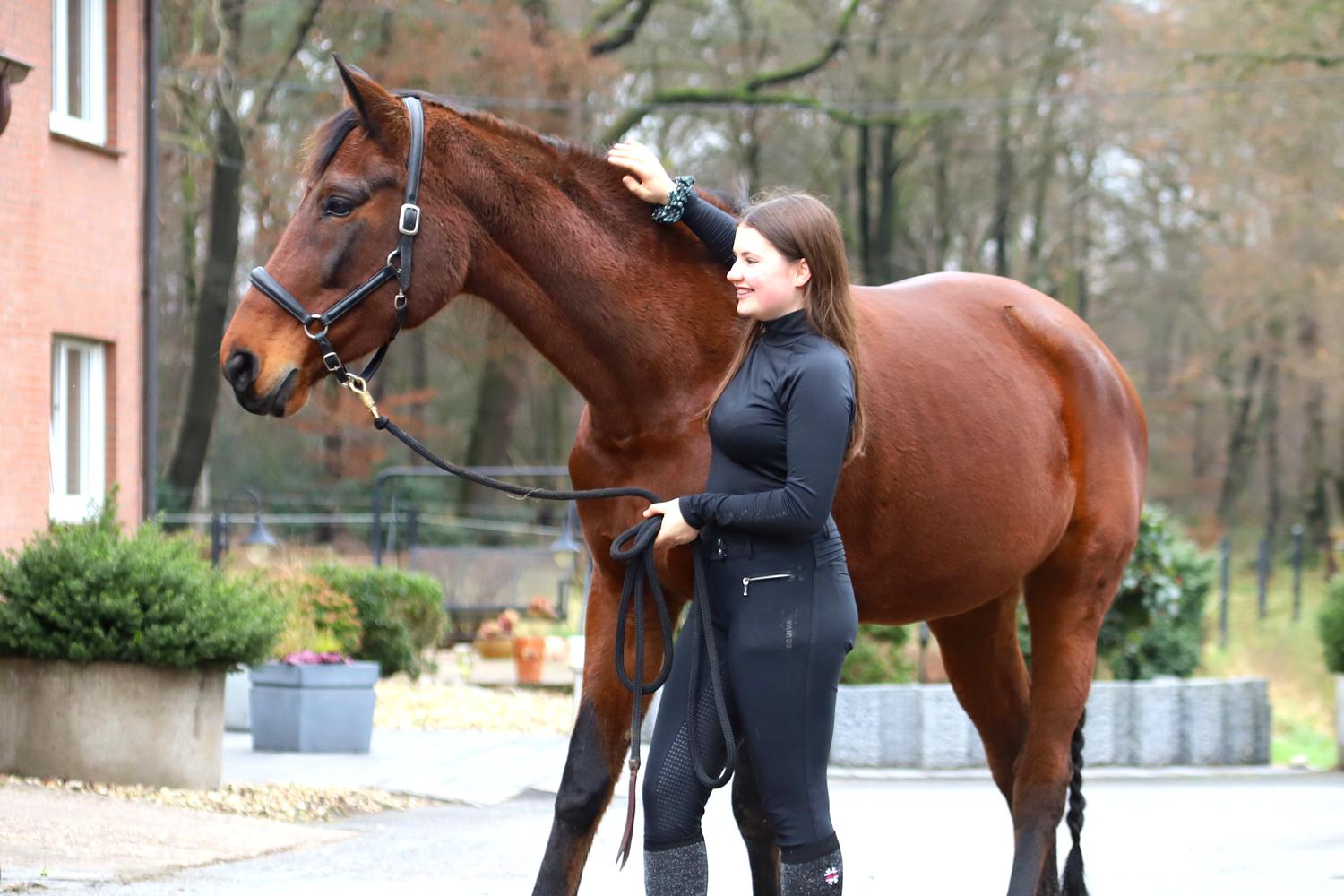 Junge Frau steht entspannt neben einem Pferd auf dem Hof